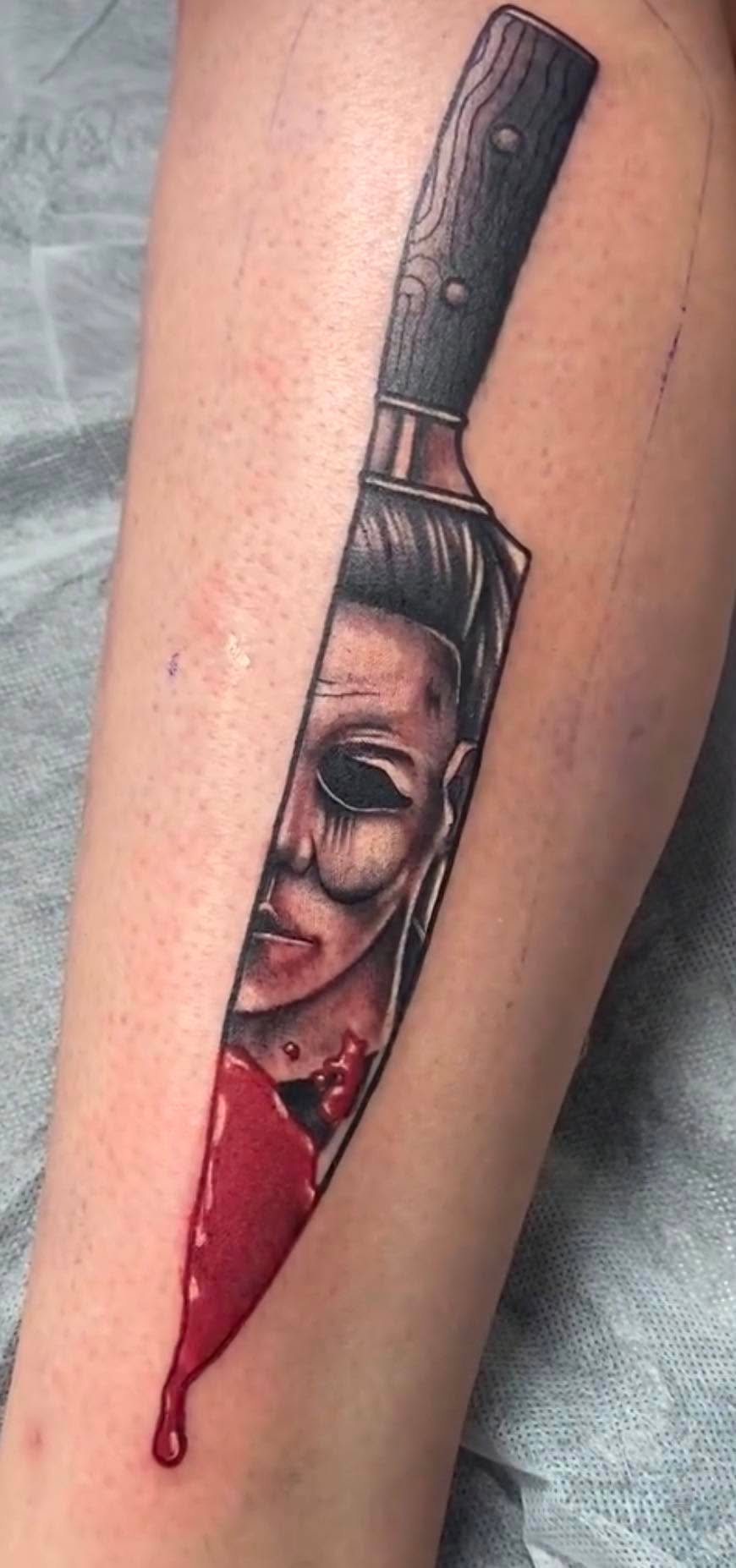 Black and grey horror movie mask in a bloodied knife tattoo by Love and Chaos Tattoo Gallery, Mudgee NSW